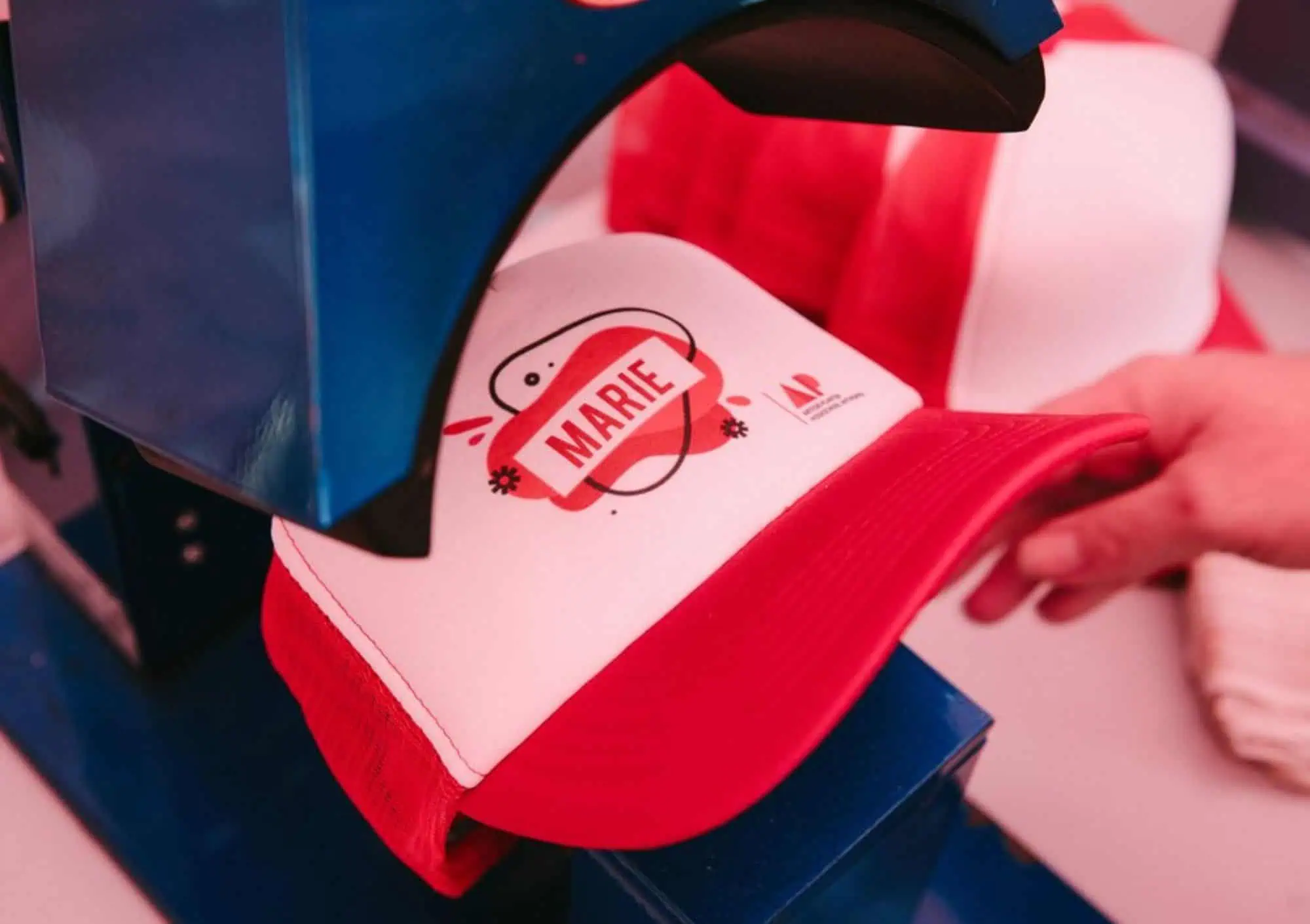 Hittepers Naam Personalisatie Close-up van een professionele blauwe hittepers die de naam 'MARIE' bedrukt op een wit-rode cap tijdens een live merkactivatie.