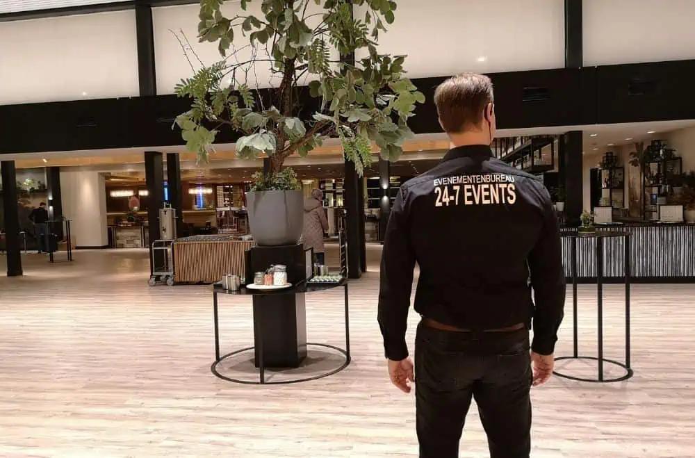 Rugfoto van een professionele eventmanager in een zwart shirt met de gele opdruk EVENEMENTENBUREAU 24-7 EVENTS. Hij overziet de ruimtelijke foyer van een congreslocatie in Ede, met blik op de zaaldoorgangen en de voorbereiding van het lunchbuffet