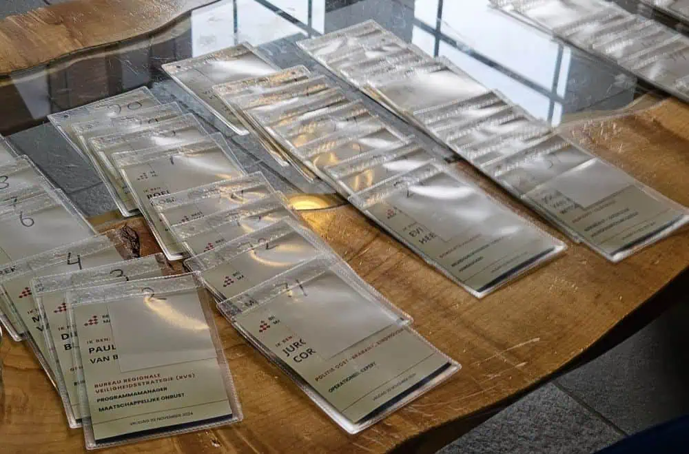 Detailopname van rijen voorbedrukte congresbadges in transparante hoezen op een houten tafel. De persoonsnamen zijn afgedekt tegen datalekken (AVG-proof), terwijl de onderzijde van elke badge het individuele zalen- en breakout-programma van de specifieke bezoeker toont.