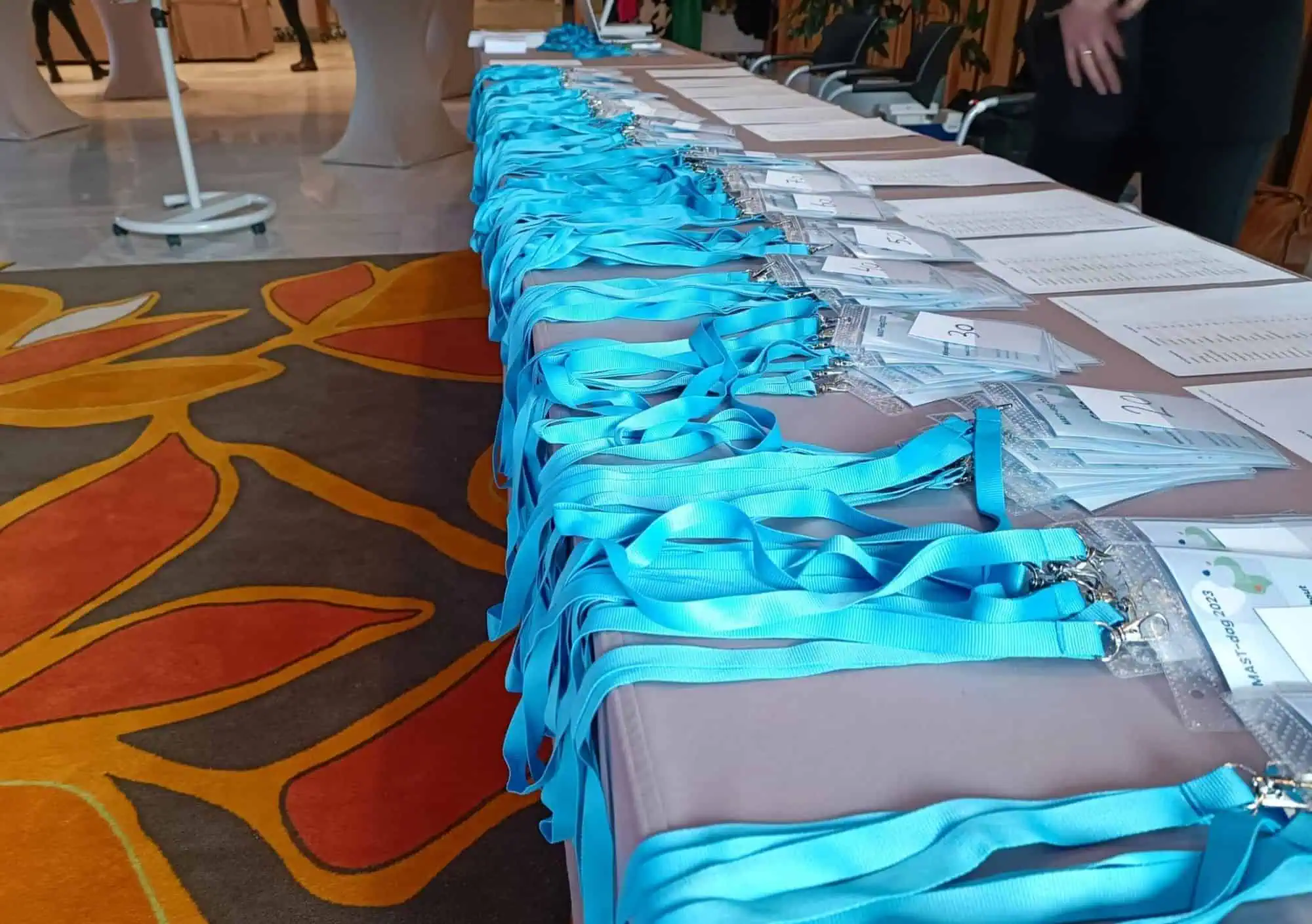 Perspectiefopname van een lange registratietafel volgelegd met felblauwe lanyards en gepersonaliseerde congresbadges in transparante hoezen, geordend op nummer. Op de achtergrond is een sfeervol tapijt met oranje bloemmotief te zien en een eventmanager die de aanmelddocumenten voorbereidt.