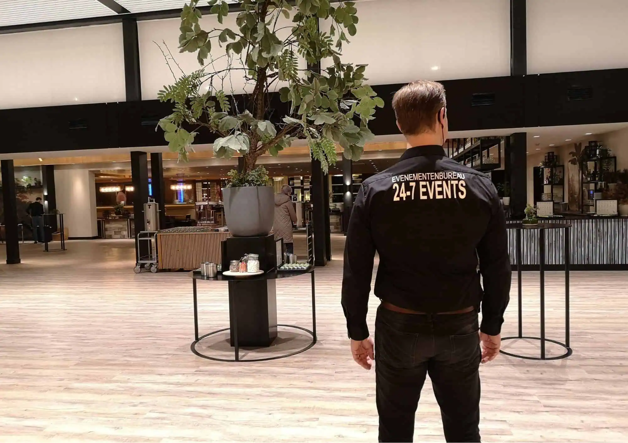 Rugfoto van een professionele mannelijke eventmanager die een zwart shirt draagt met de gele opdruk EVENEMENTENBUREAU 24-7 EVENTS. Hij overziet een moderne, luxe hotellobby met een grote centrale plantenbak op een congreslocatie.
