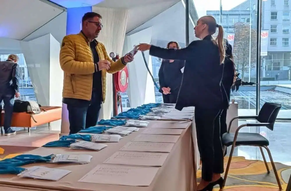 Een hospitality medewerkster van Evenementenbureau 24-7 Events reikt glimlachend een blauwe lanyard met voorbedrukte congresbadge uit aan een mannelijke bezoeker over een overzichtelijke registratietafel in een moderne, lichte foyer.