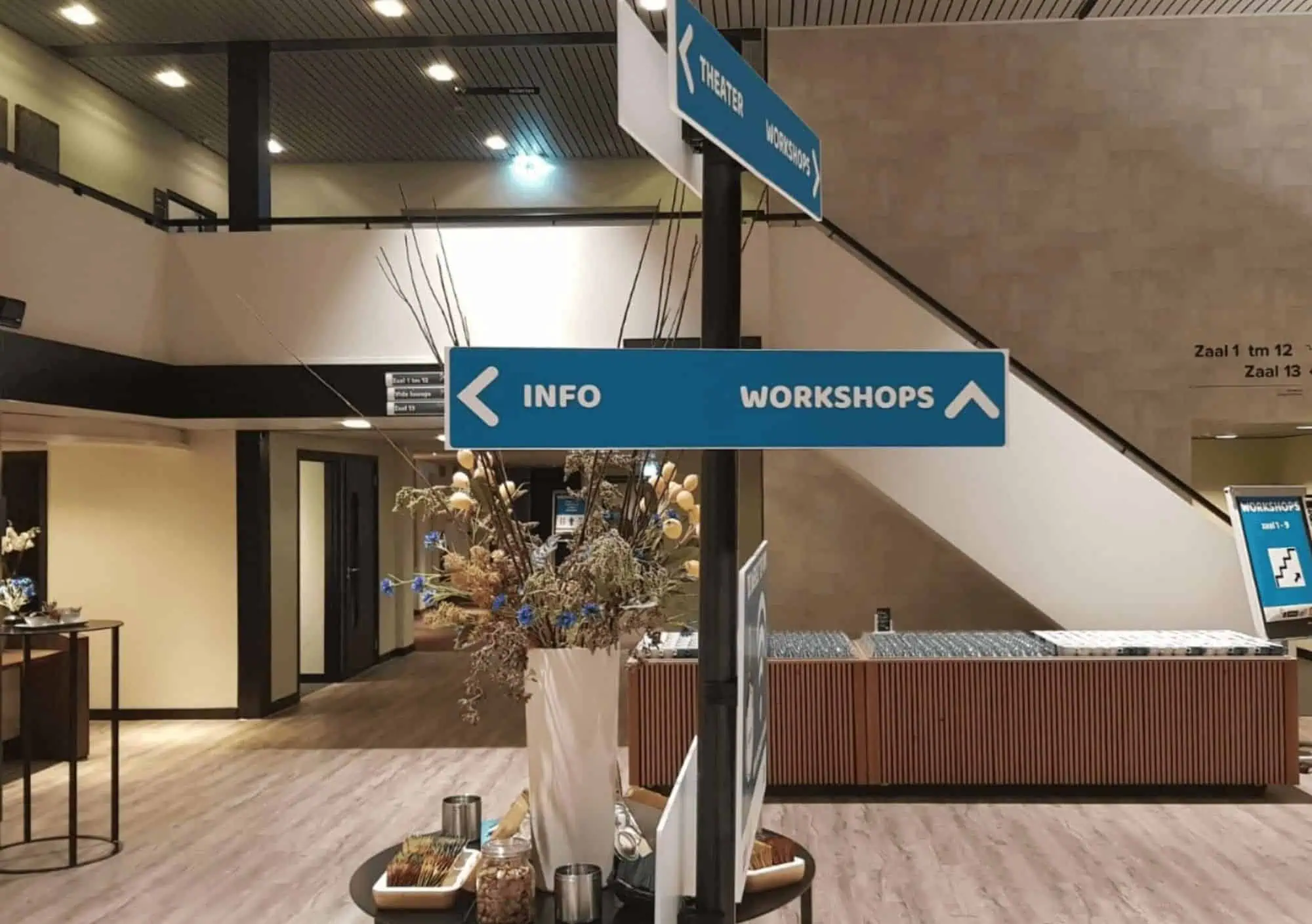 Professionele blauwe wegwijzerbordjes met witte opschriften INFO, WORKSHOPS en THEATER gemonteerd op een zwarte zuil in de centrale hal van een congreslocatie in Utrecht. Op de voorgrond staat een vaas met droogbloemen op een ronde tafel, en op de achtergrond is een ruimtelijke foyer met trap naar zalen 1 t/m 12 te zien.